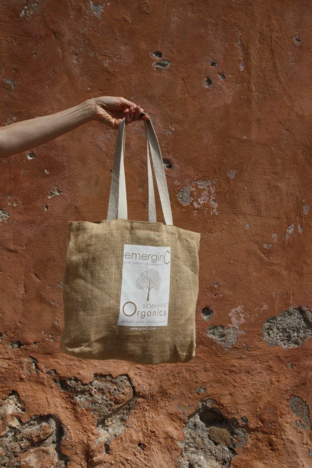 emerginC juten tas van Scientific Organics lijn die wordt vastgehouden tegen rood-bruine muur